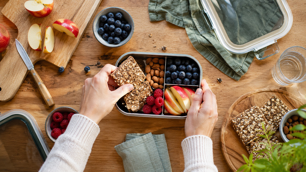 Hender legger grove knekkebrød, bær, nøtter og oppskåret frukt i en matboks på et kjøkkenbord.